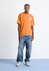 Orange bomuld T-shirt med hvid grafisk print på brystet, parret med løse blå denim jeans og grå sneakers. Glat tekstur.