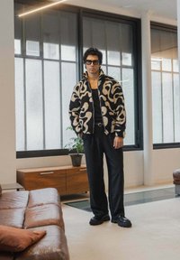 Veste à motifs noir et crème en tissu texturé, associée à un pantalon noir et des chaussures noires imposantes. Design élégant avec fermeture éclair.