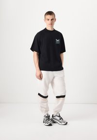 Musta ylimitoitettu t-paita, jossa on logosuunnittelu ja raglan-hihat, yhdistettynä valkoisiin jogginghousuihin, joissa on mustia ja beigen värisiä raitoja.
