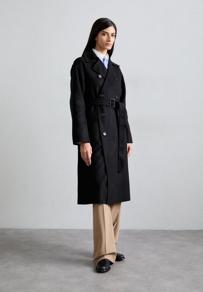 Μαύρο διπλό ραμμένο παλτό από μαλλί με ζώνη στη μέση, διακοσμημένο με μεγάλα κουμπιά και πλαϊνές τσέπες, συνδυασμένο με μπεζ παντελόνι και μαύρα παπούτσια.