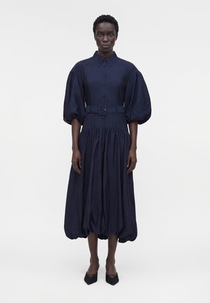 Γυναίκα που φοράει ένα μίντι φόρεμα σε μπλε ναυτικό χρώμα με ζώνη, με φουσκωτά μανίκια μέχρι τον αγκώνα και σούρα στο τελείωμα, σε συνδυασμό με μαύρες μυτερές γόβες.