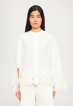 Young woman with long dark hair wearing a white button-up blouse with wide sleeves and floral lace trim at cuffs and hem.