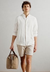 Chemise légère blanche à manches longues, short texturé beige et sac de voyage tan avec des accents marron, tenu dans la main droite.
