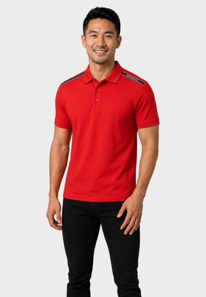 Uomo sorridente che indossa una polo rossa a maniche corte con dettagli a righe nere sulle spalle e pantaloni neri, in piedi davanti a uno sfondo semplice.