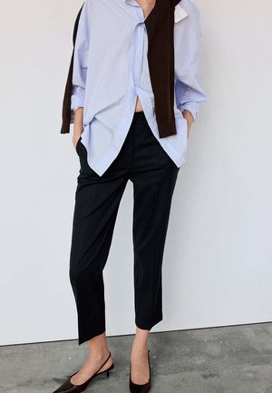 Personne portant une chemise boutonnée à rayures bleu clair, un pantalon noir court, des talons slingback marron, avec un pull foncé drapé sur les épaules.