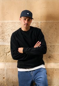 Černá mikina s texturovanou tkaninou a malým logem, zkombinovaná s modrými džíny. Námořnická kšiltovka s výrazným logem. Pozadí tvoří kamenná zeď.