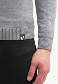 Rechte Hand liegt neben schwarzen Hosen, trägt einen grauen, gerippten Pullover mit einem kleinen schwarz-weißen Logo-Tag in der Nähe der Taille.