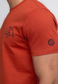 Červené bavlněné tričko s krátkým rukávem, zdobené modrým potiskem a kulatým logem na levém rukávu, hladká textura a dobře padnoucí design.