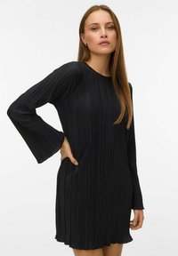 Kobieta z długimi prostymi włosami, ubrana w czarną plisowaną sukienkę z długim rękawem i szerokimi mankietami, stojąca z jedną ręką na biodrze.