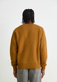Brauner Strickpullover mit lockerer Passform, gerippten Bündchen und Saum, von hinten gezeigt. Der Stoff hat eine weiche Textur und wird mit grauen Hosen kombiniert.