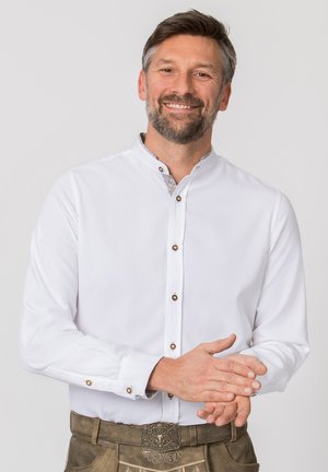 Smiling man with beard wearing white buttoned shirt and traditional brown leather pants with decorative belt buckle, hands clasped.