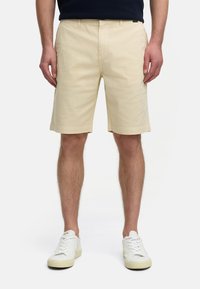 Shorts en coton beige à coupe droite, avec passants de ceinture et poches latérales, associés à des baskets blanches, présentant une texture lisse.