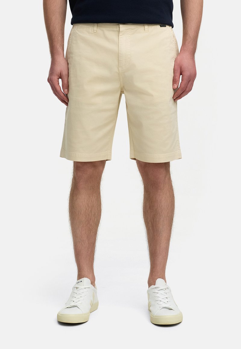 Shorts en coton beige à coupe droite, avec passants de ceinture et poches latérales, associés à des baskets blanches, présentant une texture lisse.