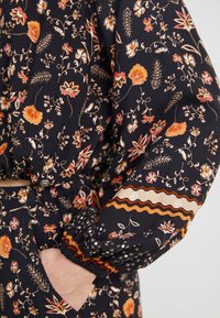 Blouse à motifs avec un fond bleu marine foncé, ornée de motifs floraux en orange, rose et crème. Manches élastiques avec des garnitures décoratives.