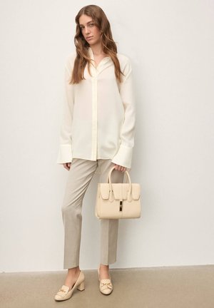 Bezs színű kézitáska strukturált kialakítással és fedeles záródással, világos krémszínű blúzzal és hozzá illő nadrággal viselve. Máglyázott díszítésű magassarkú cipő.