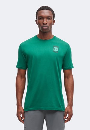 Man draagt een groen Under Armour T-shirt met korte mouwen en grijze broek, staand tegen een effen witte achtergrond.