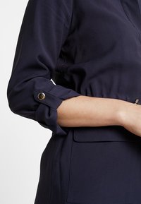 Camicia blu navy con maniche arrotolate, dettagli con bottoni e vita strutturata. Tessuto morbido con una texture liscia e dettagli con tasche laterali.