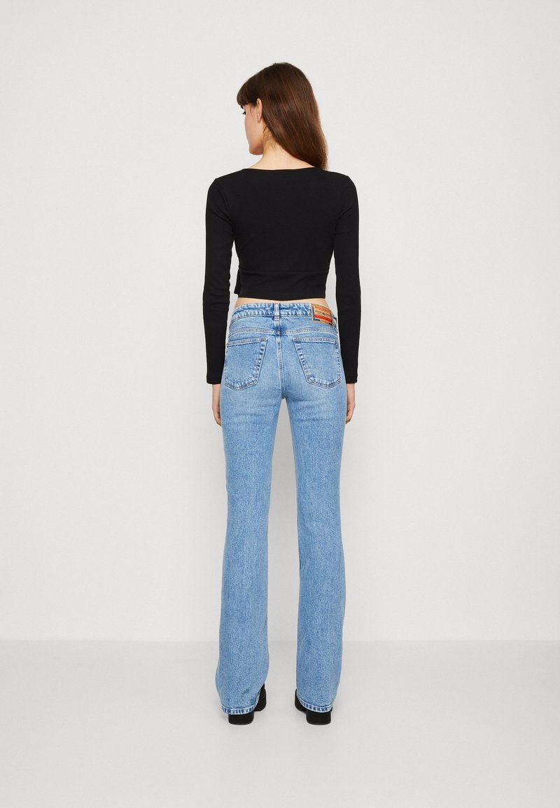 Blaue Schlagjeans mit hoher Taille, die eine dezente ausgewaschene Textur und zwei Gesäßtaschen aufweisen. Kombiniert mit einem engen schwarzen Langarmtop.