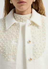 Chaqueta de lana blanca con un cuello y parte delantera adornados con perlas, botones redondos de imitación de perlas y una superficie texturizada.