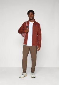 Casaco de lã castanho, t-shirt branca, calças castanhas e sapatilhas cinzentas. A figura segura uma mochila rosa e está diante de um fundo neutro.