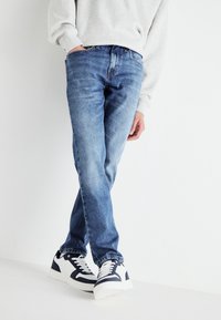 Jeans de denim azul ajustados con un acabado desgastado, con cinco bolsillos y detalles de costura, acompañados de zapatillas blancas y marinas.