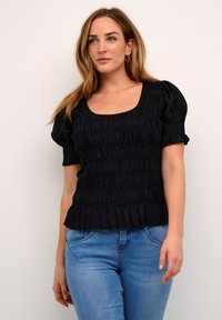 Kvinde iført en sort rynket kortærmet top og lyseblå jeans, stående imod en ensfarvet lys baggrund.