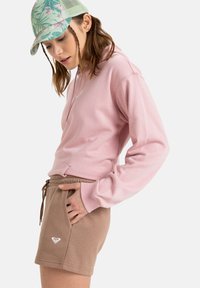 Sweat à capuche rose avec cordon de serrage et poche kangourou associé à un short marron avec taille élastique et poches latérales, détail du logo.