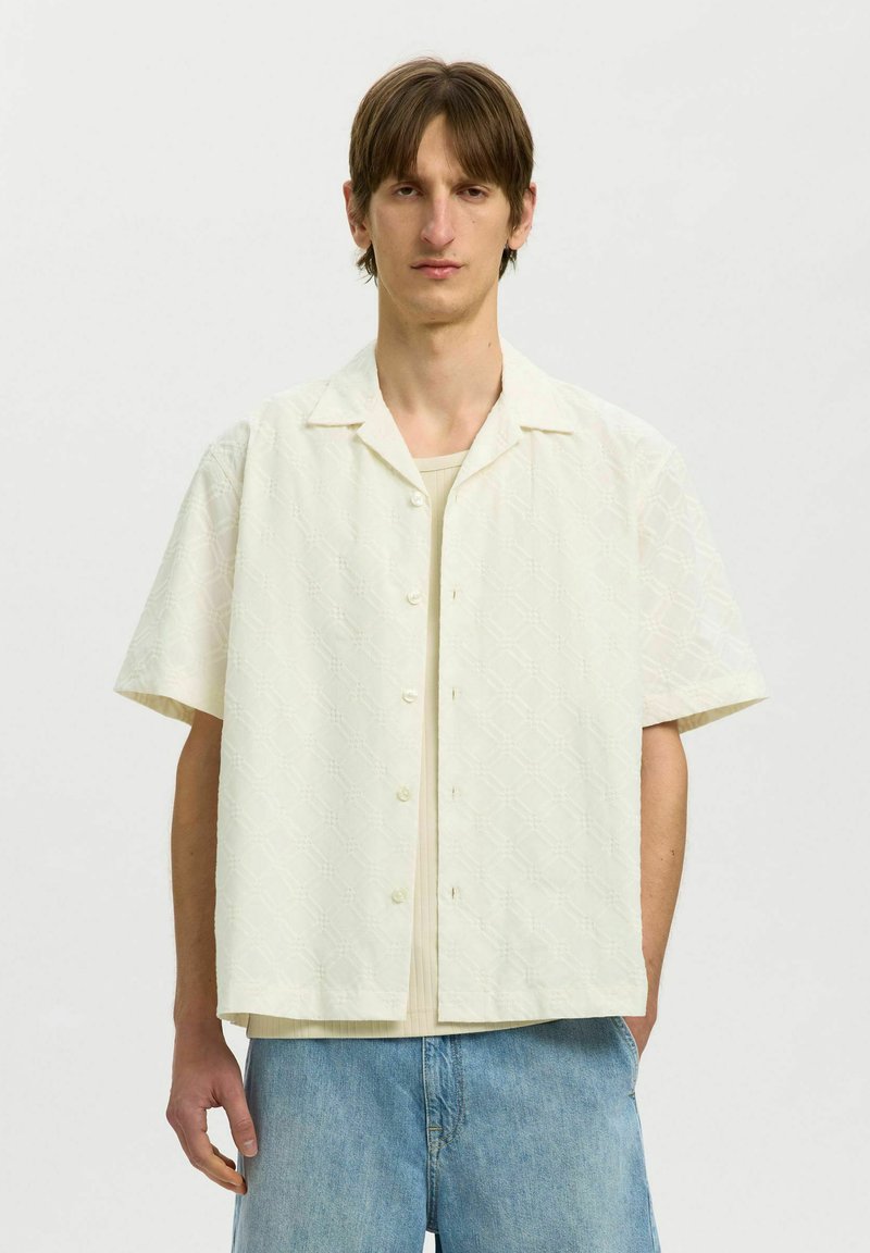 Jeune homme aux cheveux bruns portant une chemise texturée écrue à manches courtes boutonnée sur un haut beige et un jean bleu clair, debout devant un fond uni.