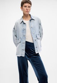 Gai zila denim jaka ar nedaudz pārlielu piegriezumu, pogu aizdare, divas priekšējās kabatas un saplēsts apakšmalas dizains. Kombinēta ar tumši ziliem samta biksēm.