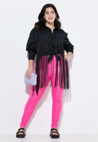 Schwarze Hemdbluse mit Fransen-Details, rosa Kunstleder-Leggings und einer helllila gesteppte Handtasche, kombiniert mit schwarzen Plateausandalen.