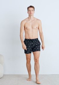 Sorte badshorts med flerfarget logo-mønster, justerbar midje og sidelommer. Modellen er uten skjorte og står på en teppebelagt overflate.