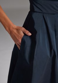 Granatowa spódnica plisowana o teksturowanej powierzchni i otwartej kieszeni. Materiał wydaje się gładki i lekkie. Ręka spoczywa na kieszeni.