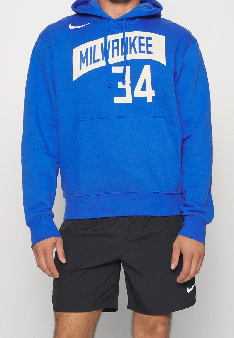 Sweat à capuche bleu avec texte blanc "MILWAUKEE 34" sur le devant, poche kangourou avant et capuche à cordon. Porté avec un short noir et un logo.