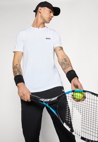 Fehér sportpóló "BOSS" logóval, fekete csuklószalag, fekete nadrág. Teniszütőt és egy zöld teniszlabdát tart a kezében.