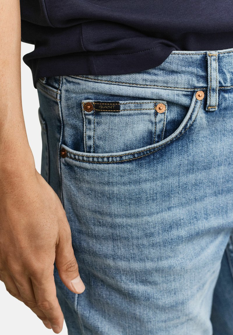 Primer plano de una persona con pantalones vaqueros de mezclilla azul claro con una pequeña etiqueta negra con el logo cerca del bolsillo delantero y una camisa oscura.