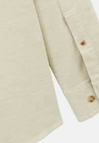 Chemise beige à côtes avec des manches longues. Caractéristiques : poignets à boutons et ourlet arrondi. La texture est douce avec des détails visibles du tissage du tissu.