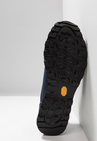 Svart gummisåle med dyp profil, med et gult Vibram-innslag. Siden viser mørkeblått materiale med en strukturell finish.