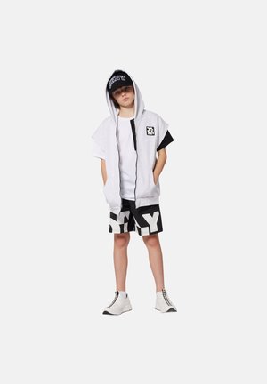 Haut sans manches gris avec des accents noirs, t-shirt blanc, shorts noirs avec de grandes lettres blanches, et chaussures blanches avec des détails noirs.