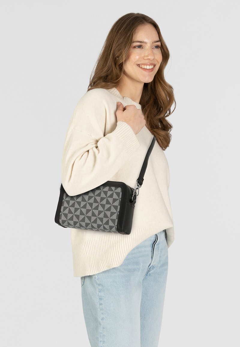 Borsa nera con un motivo geometrico grigio. Dotata di chiusura con zip e tracolla regolabile. Indossata a tracolla su un maglione chiaro.