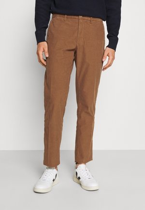 Man draagt bruine rechte broek gecombineerd met witte sneakers en een donkere longsleeve, staand tegen een effen achtergrond.