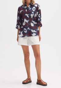 Chemise à boutons maroon et blanche avec un motif abstrait et des accents bleus, associée à un short blanc et des sandales brunes à lanières.
