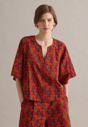 Donna con capelli corti che indossa un top e pantaloni larghi coordinati con motivo foglie rosse, arancioni, viola e verdi, in piedi con le mani nelle tasche.