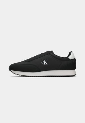 Chaussures de sport noires avec une tige en tissu lisse, des accents blancs sur le talon et une semelle blanche épaisse présentant un motif texturé. Logo sur le côté.