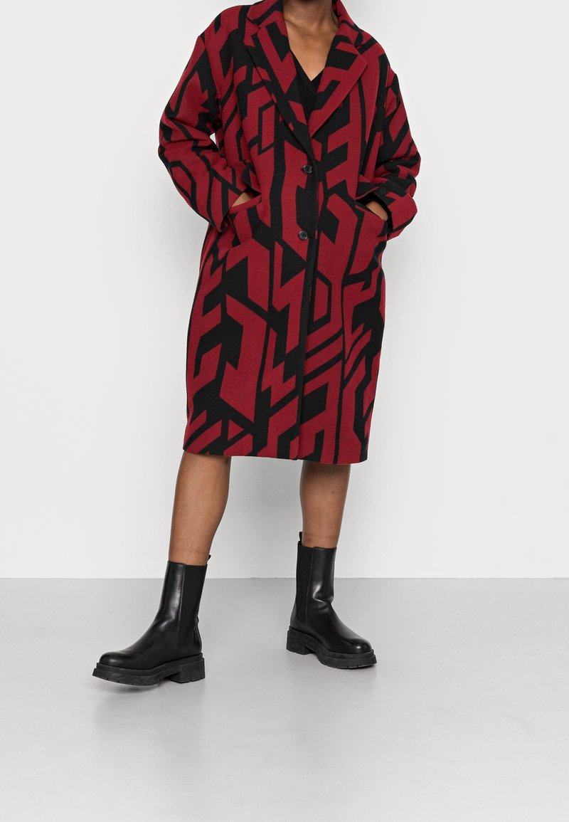 Rot-schwarz gemusterter Mantel mit lockerem Schnitt, Reverskragen und Knöpfen; getragen über einem schwarzen Outfit und kombiniert mit klobigen schwarzen Stiefeln.