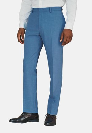 Mann trägt hellblaue Anzughose, weißes Hemd und schwarze Lederschuhe, steht vor einem schlichten weißen Hintergrund.