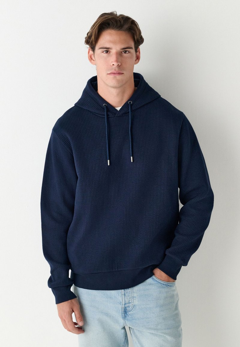 Laivastonsininen waffle-kuvioitu huppari, jossa on suuri huppu, säädettävät kiristysnauhat, resorit hihansuissa ja rento istuvuus. Yhdistetty vaaleisiin farkkuihin.