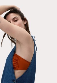 Vestido de mezclilla con escote en V y cortes laterales, que presenta un detalle de lazo en la parte posterior. Se puede ver un top de bikini naranja debajo.