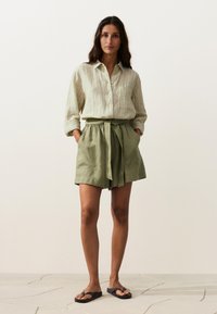 Femme debout les mains dans les poches, portant une chemise rayée beige clair boutonnée, un short vert olive ceinturé et des tongs noires.