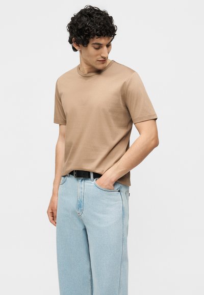 Jeune homme portant un t-shirt beige à manches courtes et un jean bleu clair, regardant vers le bas avec une main dans sa poche, sur un fond uni.