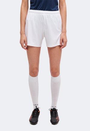 Persona che indossa pantaloncini sportivi bianchi, calzini lunghi bianchi e tacchetti neri, in piedi davanti a uno sfondo uniforme.
